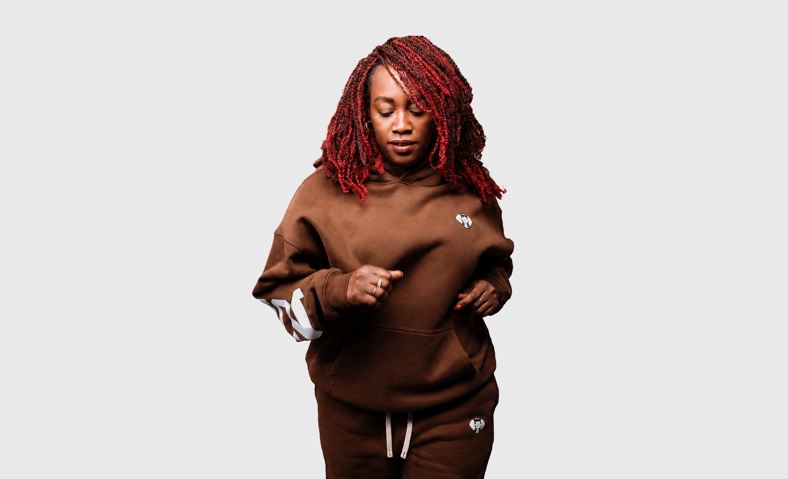 A woman with red dreadlocks wearing a brown sweatshirt and matching sweatpants with white accents, standing against a light gray background.
